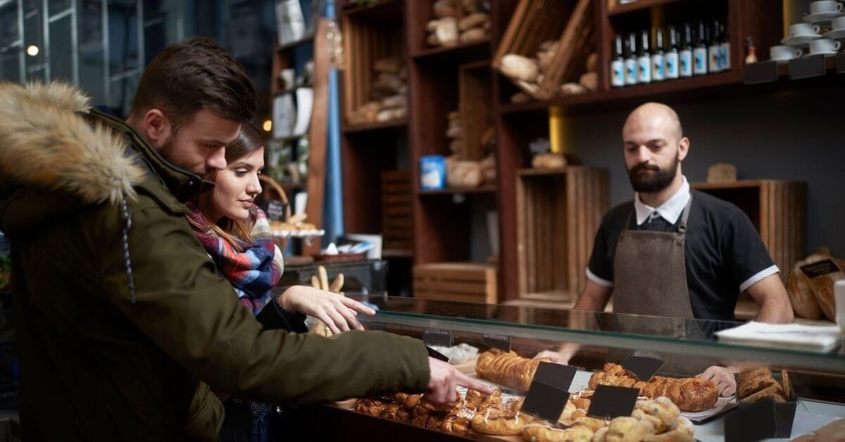 Couple shopping at a small bakery, pointing at a croissant to get the bakers attention