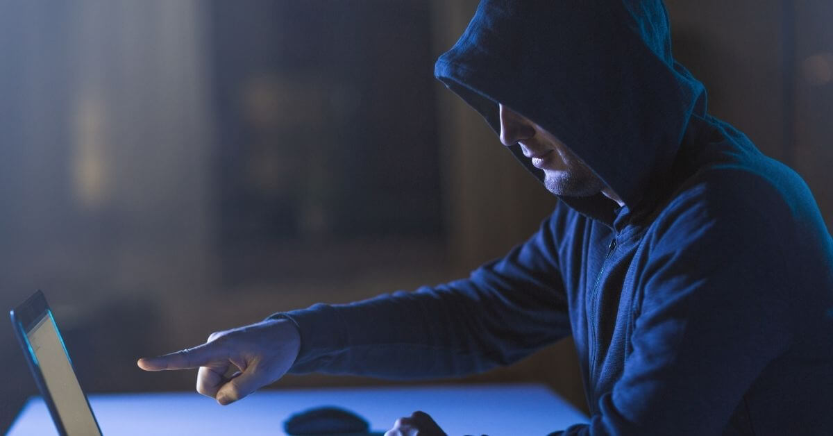 A man in a dark hoodie points at a computer in a darkened room