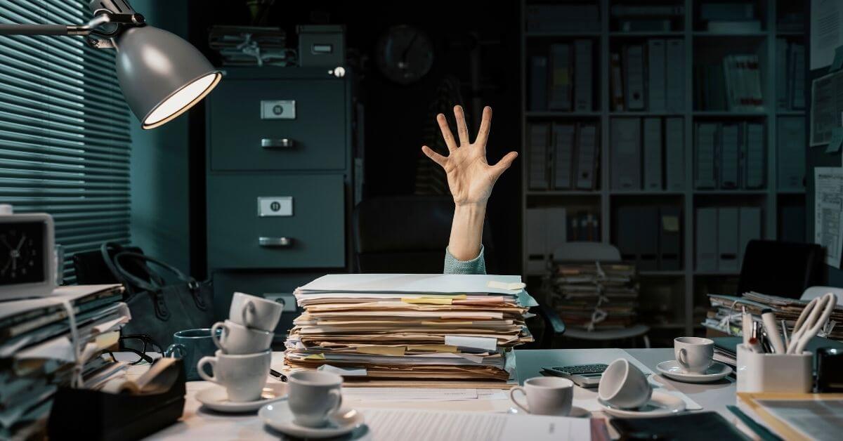 In a darkened room, illuminated by a single lamp, a hand reaches out under a desk. It is straight up, flat and open, over a parcell of papers and documents on the grey desk. Empty white cups, rounded, with smooth handles, litter the desk.
